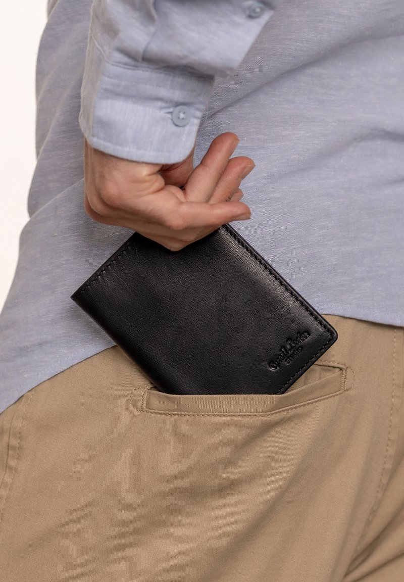 Juodas odinis piniginė, dalinai matoma kišenėje, pasižyminti glotnia tekstūra ir minimalistišku dizainu. Išorėje išspaustas logotipo detalė.