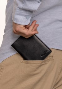 Portafoglio in pelle nera parzialmente visibile in una tasca, caratterizzato da una texture liscia e un design minimalista. Dettaglio del logo impresso all'esterno.