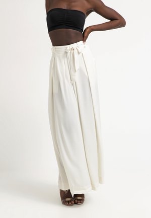 Pantalons larges blancs à taille haute, avec des plis avant et une ceinture nouée. Tissu lisse avec un léger éclat. Talons bruns à lanières inclus.