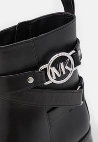 Bottine en cuir noir avec boucle circulaire argentée à logo Michael Kors et détails de sangle, gros plan sur le côté supérieur et les coutures.