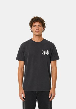 Hombre con cabello rizado y castaño vistiendo una camiseta lisa de color gris oscuro con un pequeño logo blanco de "DEUS" en el pecho izquierdo, de pie contra un fondo claro.