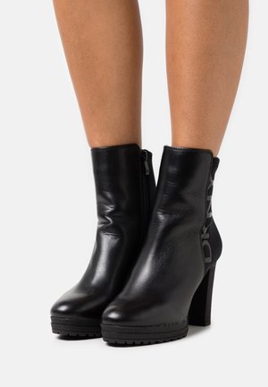 Bottes noires pour cheville en cuir brillant, avec un talon haut épais, une fermeture éclair latérale et une plateforme noire texturée avec un détail de marque.