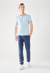 Lys blå polo skjorte med rød kant, kombinert med marineblå bukser. Modellen har på seg hvite sneakers og poserer foran en nøytral bakgrunn.