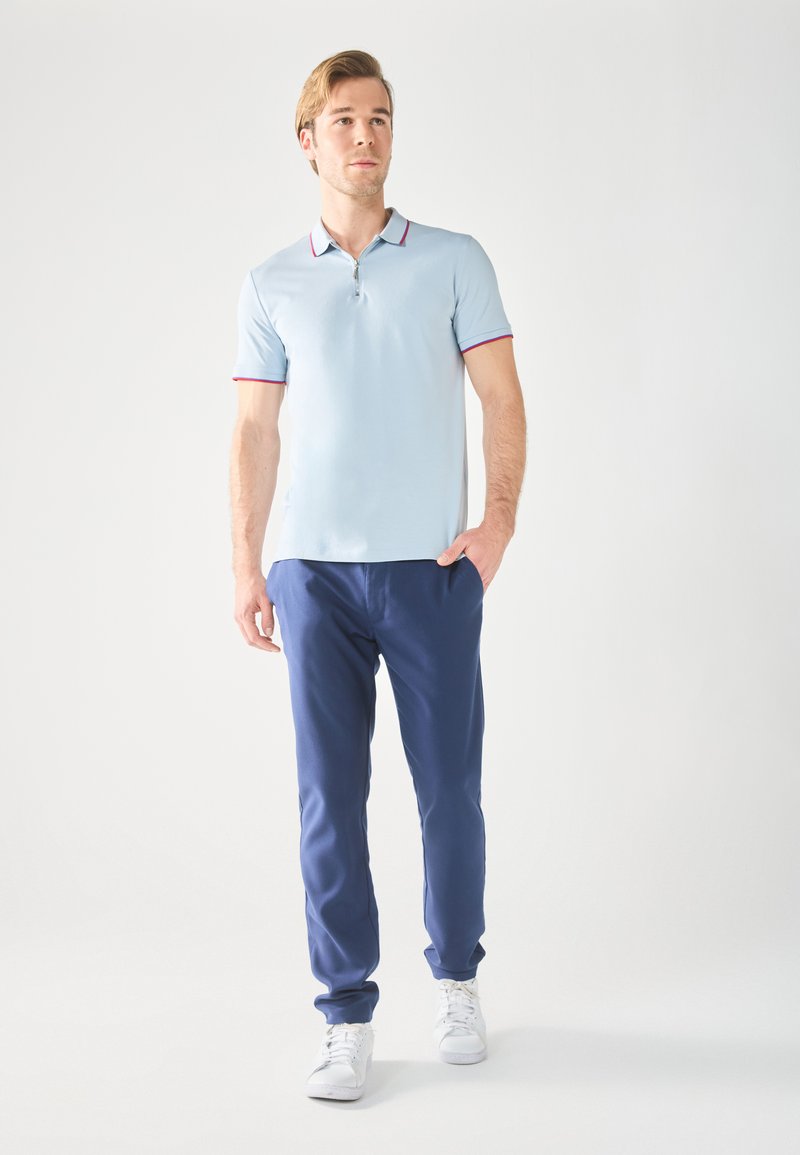 Lys blå polo skjorte med rød kant, kombinert med marineblå bukser. Modellen har på seg hvite sneakers og poserer foran en nøytral bakgrunn.