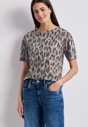 Mujer que lleva una camiseta de manga corta con estampado de leopardo en beige y negro, metida dentro de unos vaqueros azules, de pie con una mano en el bolsillo.