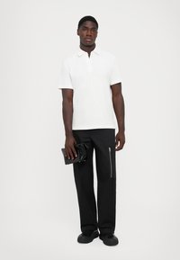 Hvit polo skjorte med korte ermer, kombinert med svarte bukser med en glidelåslomme, holdt med en svart fold-over clutch, iført svarte sko.