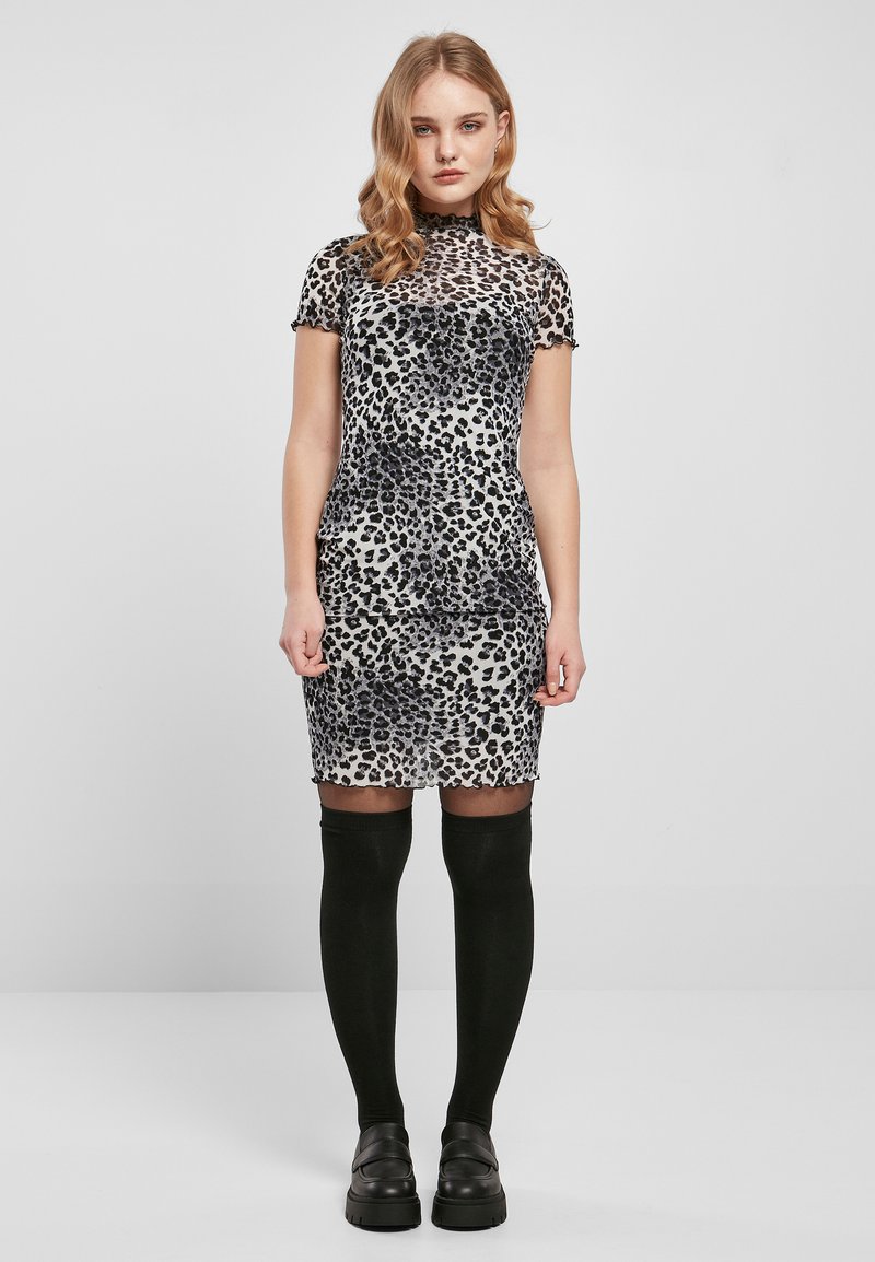 Durchsichtiges Kleid mit Leopardenmuster, kurzen Ärmeln, rundem Ausschnitt und fransigen Kanten; kombiniert mit schwarzen Strümpfen über dem Knie und klobigen schwarzen Schuhen.