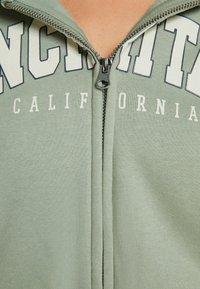 Primer plano de una sudadera verde con media cremallera que tiene la palabra "CALIFORNIA" en letras grandes con contorno blanco en el pecho.