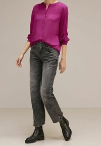 Blusa di seta magenta con bottoni sul davanti, abbinata a jeans neri dritti sbiaditi e stivali neri alla caviglia, su uno sfondo neutro.
