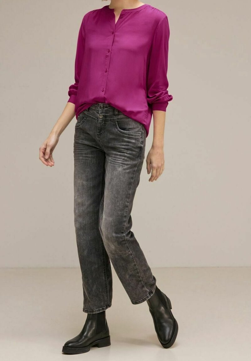 Blusa di seta magenta con bottoni sul davanti, abbinata a jeans neri dritti sbiaditi e stivali neri alla caviglia, su uno sfondo neutro.