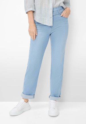 Kvinde iført lyseblå opkrogede jeans, hvide sneakers og en lysefarvet, stribet skjorte med knapper. Den ene hånd i lommen, den anden afslappet ved siden.