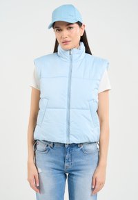 Šviesiai mėlynas be rankovių pūkinis liemenė su aukštu kaklu, priekiniu užtrauktuku ir dviem šoninėmis kišenėmis; dėvimas virš baltos marškinėlių ir derinamas su mėlynomis džinsais.