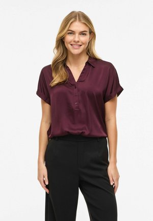 Femme souriante aux longs cheveux blonds, portant un chemisier bordeaux à manches courtes et un pantalon noir, debout devant un fond blanc uni.