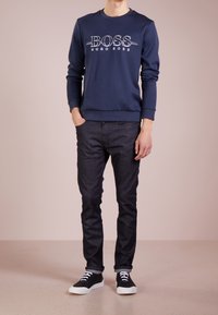 Marinblå sweatshirt med vit "BOSS HUGO BOSS" logotyp, kombinerad med mörka jeans och svarta sneakers. Slät textur och avslappnad passform.