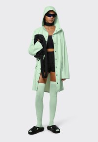 Mintgroene oversized regenjas met capuchon, zwarte crop top, zwarte shorts, kniehoge mintkleurige sokken en zwarte sandalen. Houdt een zwarte tas vast.