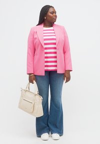 Γυναίκα που φοράει ροζ σακάκι, ριγέ πουκάμισο, τζιν παντελόνι με φαρδιά μανίκια, λευκά αθλητικά παπούτσια, κρατώντας μπεζ τσάντα, κοιτάζοντας πλάγια σε απλό φόντο.