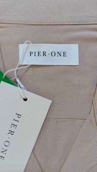 Bēšas krāsas audums ar izšūtu tekstūru, ar baltu uzlīmi, uz kuras rakstīts "PIER•ONE" un etiķeti ar apaļu caurumu.
