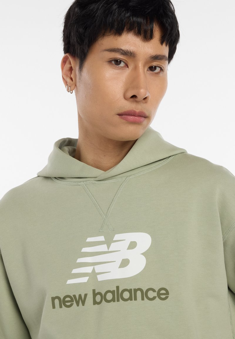 Sweat à capuche vert clair avec une poche kangourou à l'avant, une capuche à cordon de serrage, et un logo "New Balance" blanc ainsi qu'un texte imprimés de manière visible.