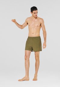 Pantaloni da bagno da uomo color verde oliva con un motivo geometrico, vita elastica e lunghezza fino a metà coscia. Modello in piedi a piedi nudi contro uno sfondo semplice.