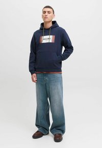 Noor mees seisab, seljas tumesinine kapuutsiga jakk "Jack & Jones" logoga, laiad helesinised teksad ja pruunid kingad, ühtlasel valgel taustal.
