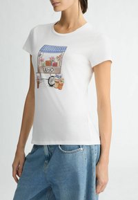 T-shirt blanc présentant un design imprimé d'une charrette à fleurs avec des pots et des fleurs. Manches courtes et silhouette ajustée.