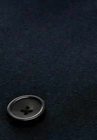Bottone nero con superficie liscia e quattro fori, appoggiato su un tessuto di feltro blu scuro con un aspetto texture.