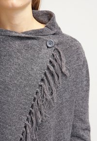 Šedý pletený šál s texturedním povrchem, má třásně na jedné straně a zapínání na knoflík u límce.