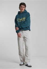Grøn hættetrøje med "Prohibite" logo i gult, hvide bukser, grå og hvide sneakers, afslappet pasform, bomuldsstof, blød tekstur.