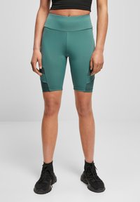 Egy nő teal színű, combközépig érő sportnadrágot visel, fekete oldal zsebekkel és fekete hálós betétekkel, fekete, zokniszerű sportcipővel párosítva.