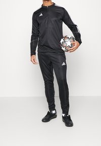 Musta Adidas-urheilupuku valkoisilla raidoilla, jossa on vetoketjullinen kaulus, kapeat housut ja teksturoitu jalkapallo. Mustat nappikengät jalassa.