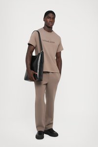 Beige t-skjorte med kort erme og preget logo, paret med matchende beige joggebukser. Svart lær skulderveske og svarte joggesko.
