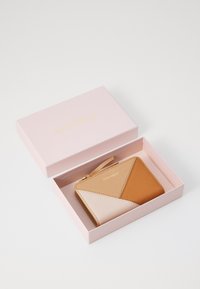 Lys brun og naken lær lommebok med geometrisk design og glidelås, presentert i en lyserosa boks med "Coccinelle" i gullbokstaver.