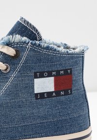 Denimové vysoké tenisky s třepenými okraji, béžovými tkaničkami a značkovým logem v červené, bílé a modré. Viditelná gumová podrážka.