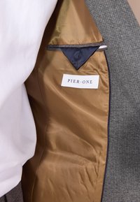 Grå blazerinnside med en glatt, gyllen fôr. Den har en marineblå lomme og en hvit etikett med teksten "PIER·ONE."
