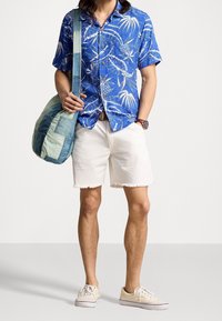 Niebieska hawajska koszula z krótkim rękawem, z białym nadrukiem palm i ananasów, w połączeniu z białymi, postrzępionymi szortami i beżowymi trampkami. Denimowa torba na ramię.