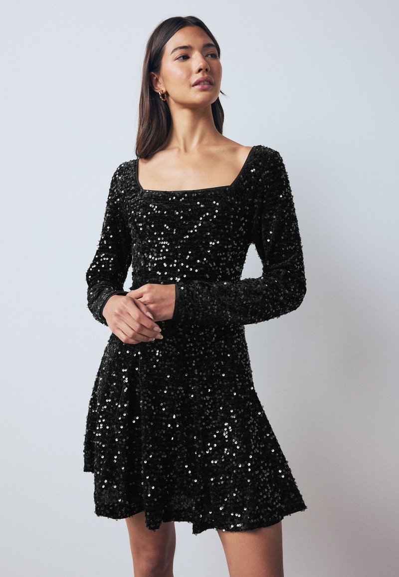 Robe en sequins noirs avec un décolleté carré, des manches longues et une jupe évasée. La robe présente une surface brillante et texturée.