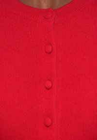 Červený pletený kardigán so zaobleným výstrihom a tromi zodpovedajúcimi gombíkmi na prednej strane. Mäkko textúrovaný s priliehavým strihom.