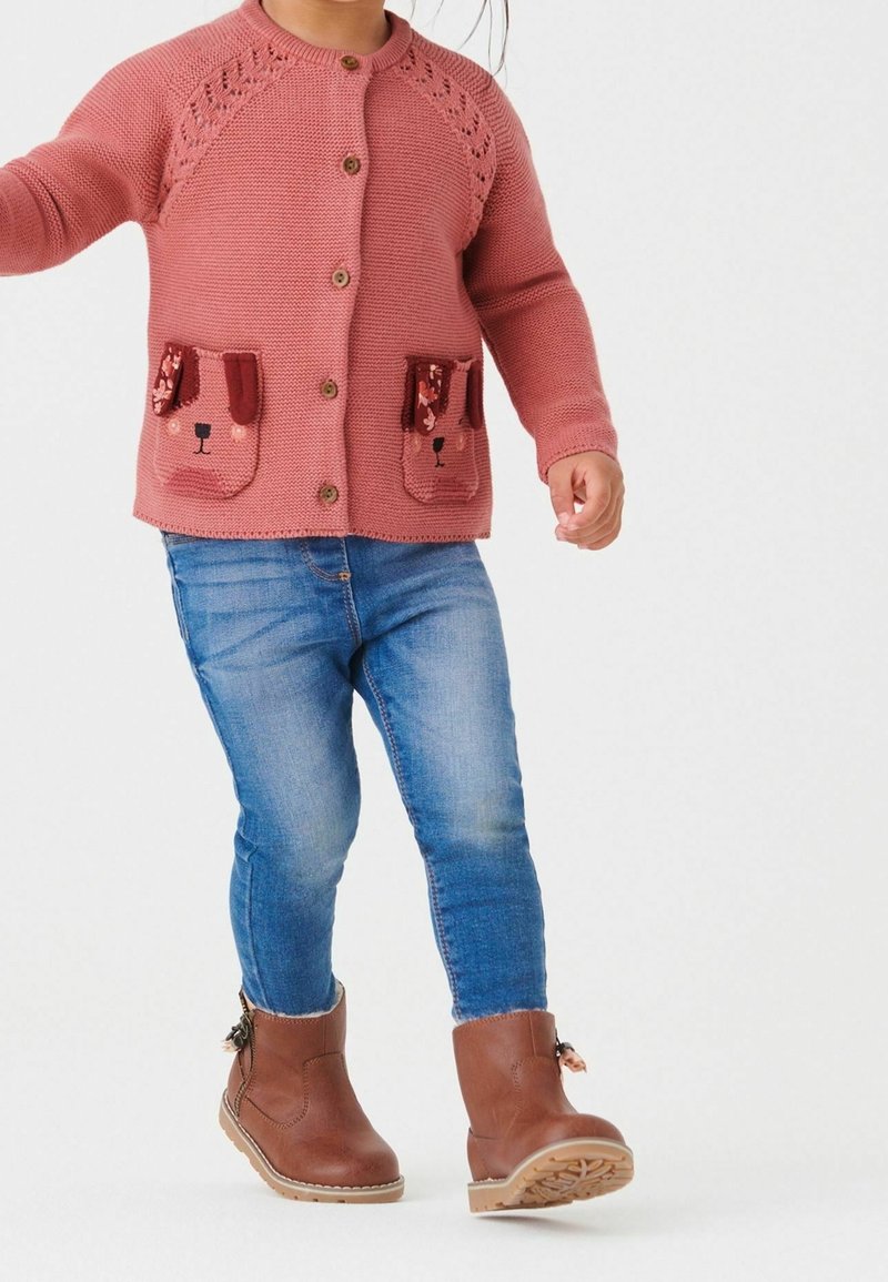 Tout-petit portant un cardigan en maille rose avec des poches à patch ours, un jean bleu et des bottes en cuir marron, marchant sur un fond blanc.