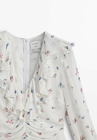 Robe blanche à fleurs avec un col en volant et des manches longues, ornée de petites fleurs multicolores et d'une fermeture éclair au dos.