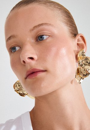 Mladá žena s uhladenými vlasmi stiahnutými dozadu, nosiaca veľké nepravidelne tvarované kladivom opracované zlaté náušnice a biely top na jednofarebnom pozadí.