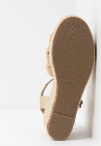 Semelle d'une sandale compensée beige avec semelle inférieure en caoutchouc texturé et détail de bride tressée, inclinée sur un fond blanc.