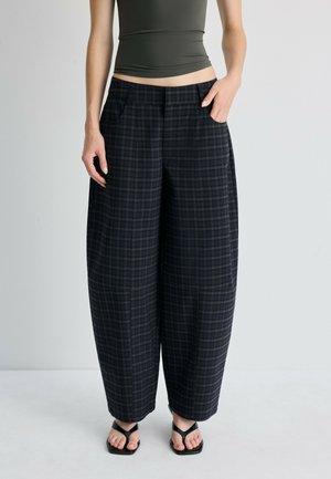 Personne portant un pantalon large à carreaux noir et vert foncé, un débardeur noir et des sandales noires à bout ouvert, debout devant un fond uni.