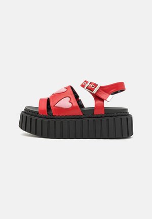 Rød platformsandal med tyk sort sål, lyserøde hjertepatches på stropperne og justerbar sølvspænde på ankelstroppen.