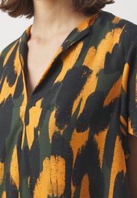 Tričko s krátkým rukávem s abstraktním vzorem v zelené, černé a oranžové barvě, s výstřihem do V a texturovanou tkaninou.