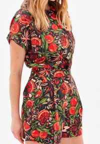 Mono corto de botones con estampado floral y mangas cortas. Presenta un vibrante estampado en naranja y verde sobre un fondo negro. Tejido texturizado con bolsillos.