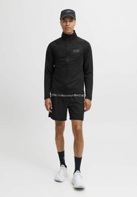 Chaqueta negra con cremallera y logo, camisa gris de patrón debajo, pantalones cortos negros, calcetines negros y zapatillas deportivas blancas. Fondo simple.