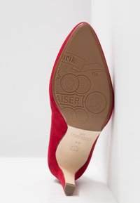 Rød ruskinds højhælet sko med spids tå. Sålen har prægede tekster og møntdesigns i en tan farve, der viser mærkets detaljer.