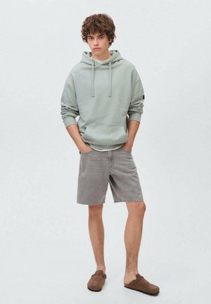 Lichtgroene hoodie met grote voorzak en trekkoord, gecombineerd met grijze denim shorts. Model draagt bruine instapschoenen.