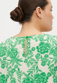 Blommig blus med grön mönstring på en krämfärgad bakgrund, nyckelhålsstängning baktill med matchande knapp och korta ärmar.