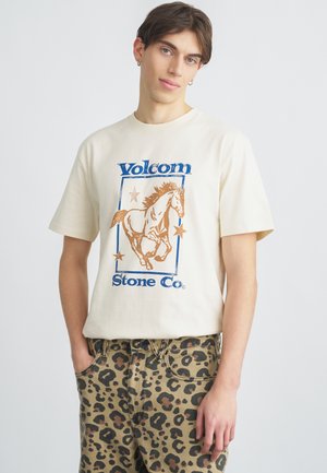 Εκρού κοντομάνικο μπλουζάκι με μπλε και καφέ γραφικό άλογο, κείμενο "Volcom Stone Co" πλαισιωμένο από μπλε πλαίσιο, συνδυασμένο με καφέ παντελόνι με λεοπάρ μοτίβο.